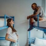 Two young women engaging in conversation in a casual hostel setting with blue bunk beds.