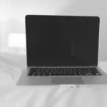 A laptop sits on a bed in a minimalist white room, perfect for remote work inspiration.
