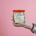A hand holding a glass jar filled with dollar bills labeled 'Where to next?' against a pink background, symbolizing savings and future planning.