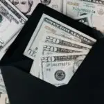 From above of dollar bills in opened black envelope placed on stack of United states cash money as concept of personal income